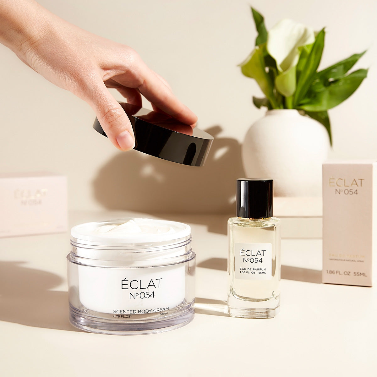 A hand is opening the lid of a jar of ÉCLAT No. 054 scented body cream, placed next to a bottle of the matching Eau de Parfum for Her. In the background, two perfume boxes and a white vase with fresh green leaves and white flowers add a clean, elegant atmosphere.