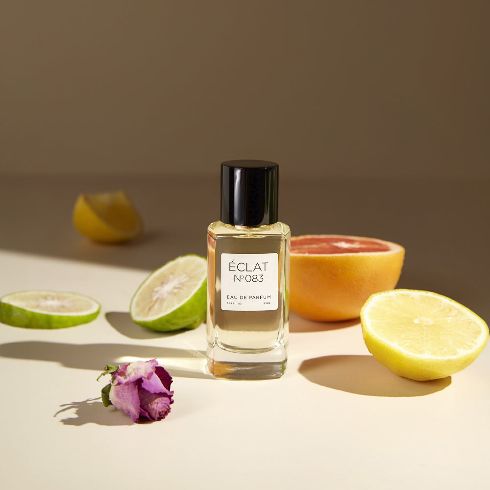 A bottle of ÉCLAT No. 083 women's perfume is placed on a soft beige surface surrounded by citrus fruits and a purple rosebud. The bright slices of lemon, grapefruit, and bergamot hint at the fragrance’s zesty and floral character.