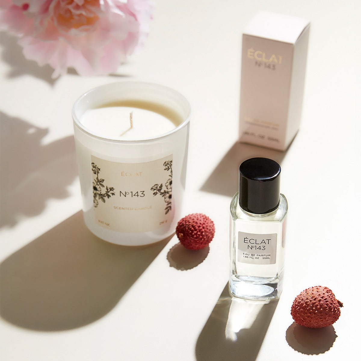 ÉCLAT Clean Rose perfume and ÉCLAT No. 143  scented candle set, displayed with lychee fruits on a light surface. The perfume bottle features a black cap and white label, while the matching candle is housed in frosted glass with floral design elements. A matching perfume box is in the background.