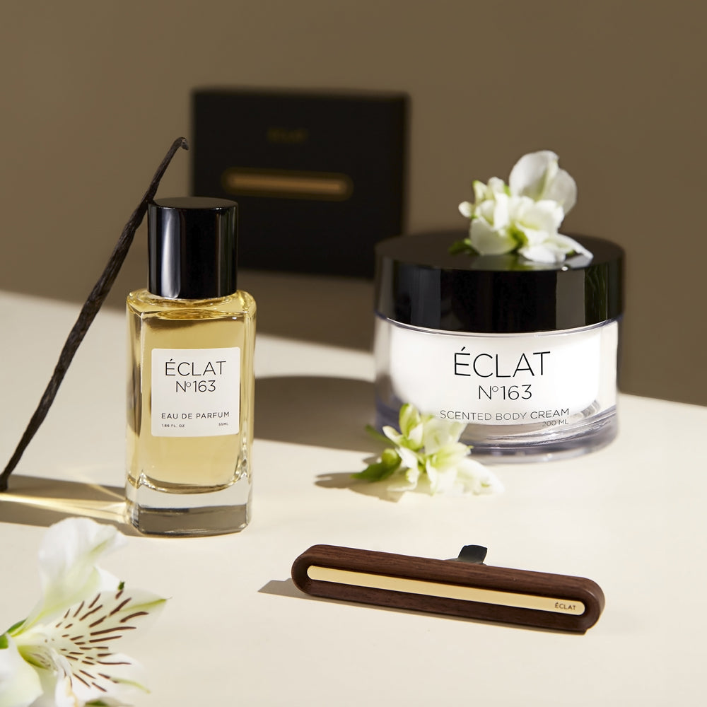ÉCLAT No. 163 women's fragrance collection, including Eau de Parfum, scented body cream, and a wooden car scent diffuser, styled with white blossoms and vanilla bean for a soft, elegant presentation on a creamy beige surface.