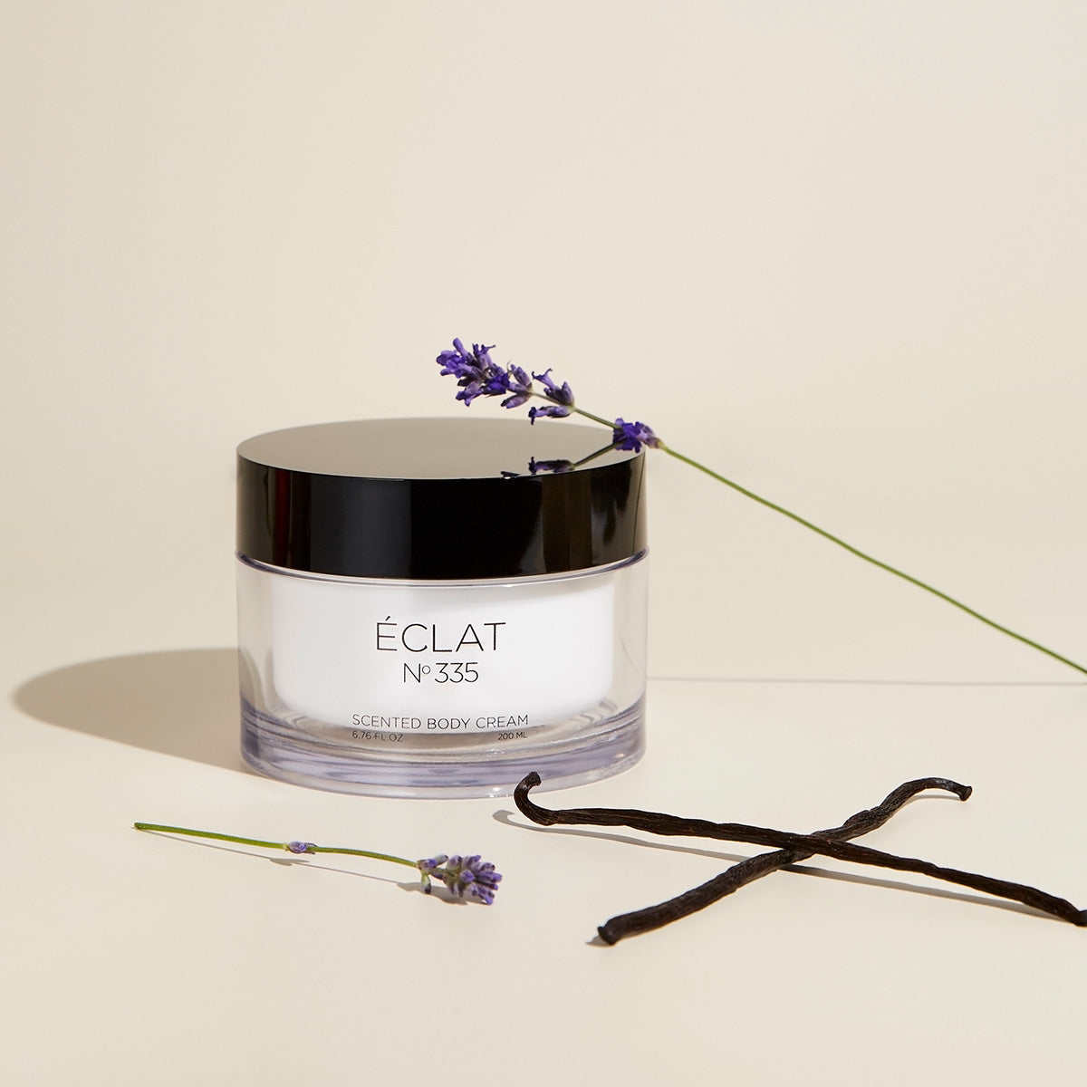 La crema corpo ÉCLAT 335 in un barattolo trasparente con coperchio nero, circondata da rametti di lavanda e baccelli di vaniglia.