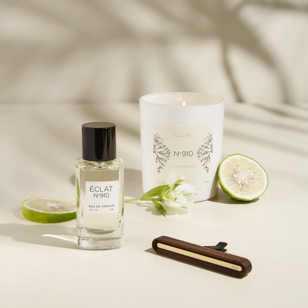 ÉCLAT No. 910 perfume bottle, matching scented candle, and car fragrance displayed together on a light surface. Decorated with bergamot slices and white blossoms, the scene highlights the fragrance trio's fresh and floral notes.