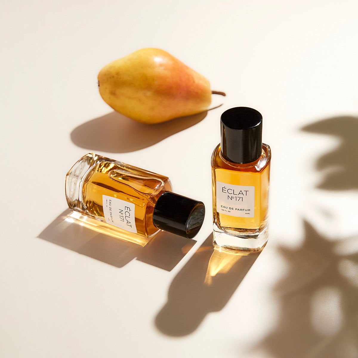 ÉCLAT Blackcurrant & Praline in eleganti flaconi di vetro con tappi neri, accanto a una pera matura, che proiettano ombre delicate.
