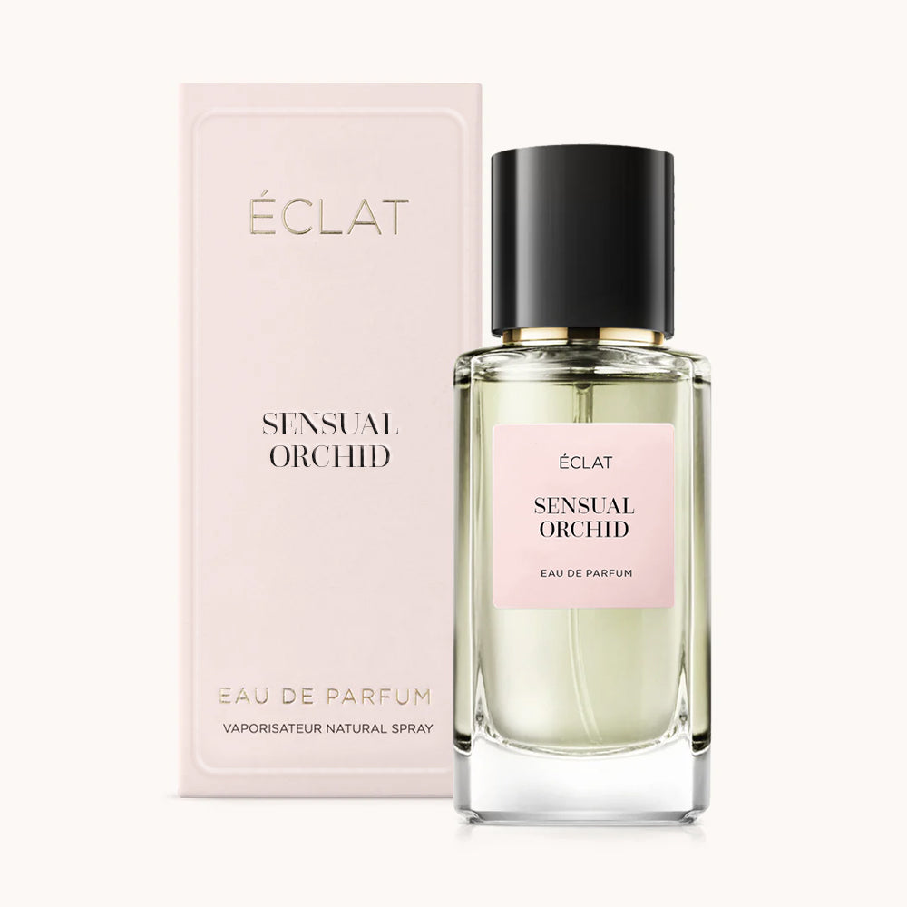 Un flacone di vetro sfaccettato con tappo nero e etichetta rosa accanto alla sua scatola di presentazione rosa per ÉCLAT Sensual Orchid.