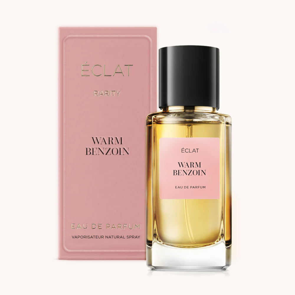 Il profumo ÉCLAT Warm Benzoin è esposto con una bottiglia di vetro trasparente con tappo nero e etichetta rosa; accanto, una scatola rosa coordinata.