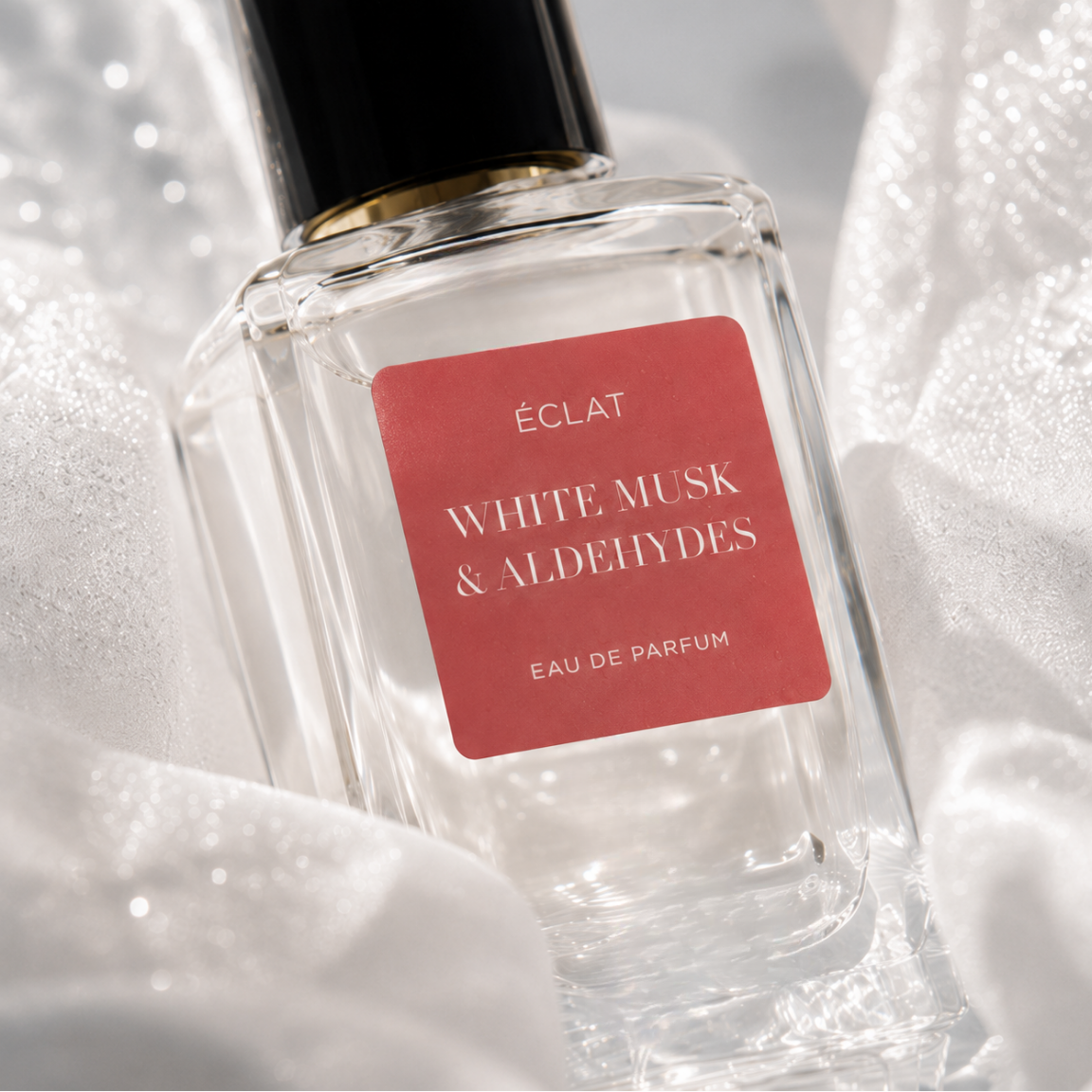 Un flacone in vetro sfaccettato di ÉCLAT White Musk & Aldehydes con etichetta rossa e tappo nero, circondato da delicato tessuto bianco.