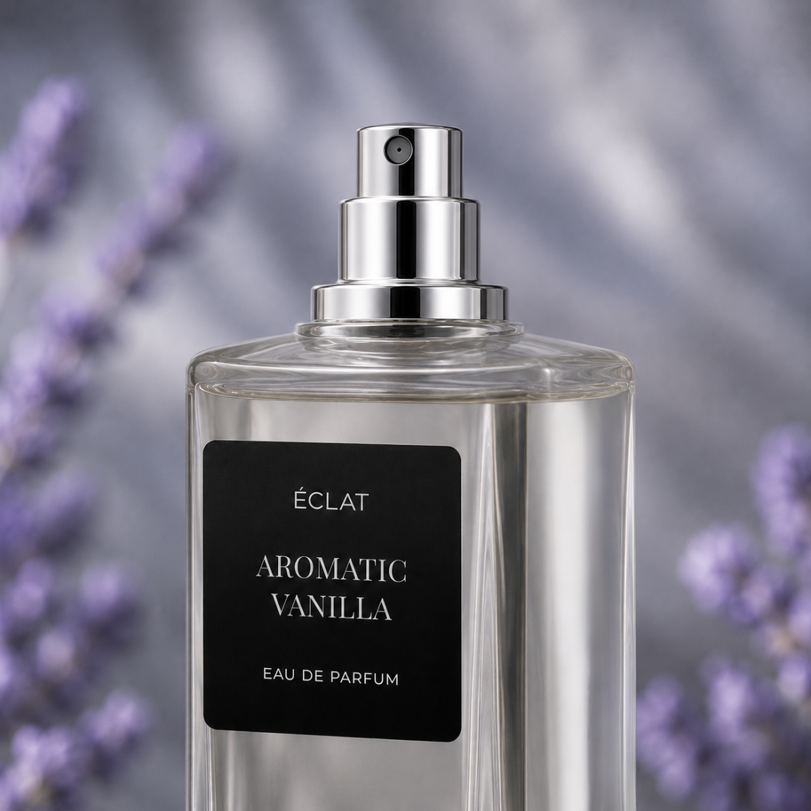 Una bottiglia di vetro trasparente di ÉCLAT Aromatic Vanilla con tappo spray argento lucido e etichetta nera opaca, su sfondo sfocato di lavanda.