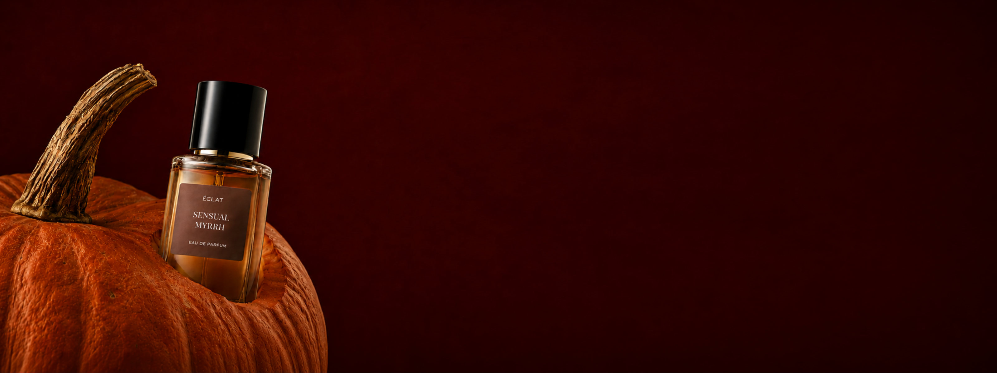 Un flacone in vetro sfaccettato del profumo ÉCLAT Sensual Myrrh è elegantemente posizionato su una zucca su uno sfondo rosso scuro intenso.