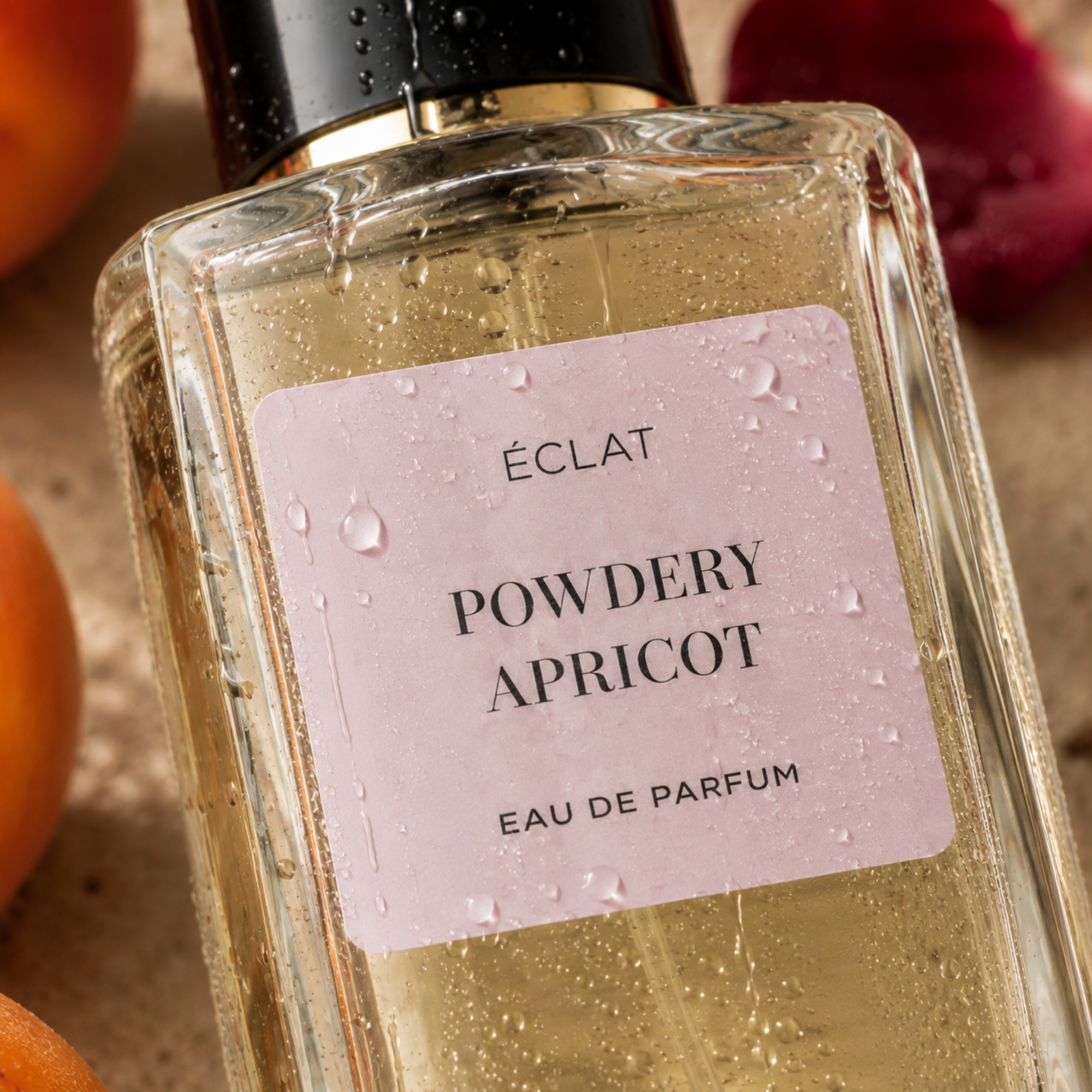 Flacone di vetro sfaccettato di ÉCLAT Powdery Apricot con etichetta rosa chiaro e tappo nero, circondato da albicocche e petali rossi.