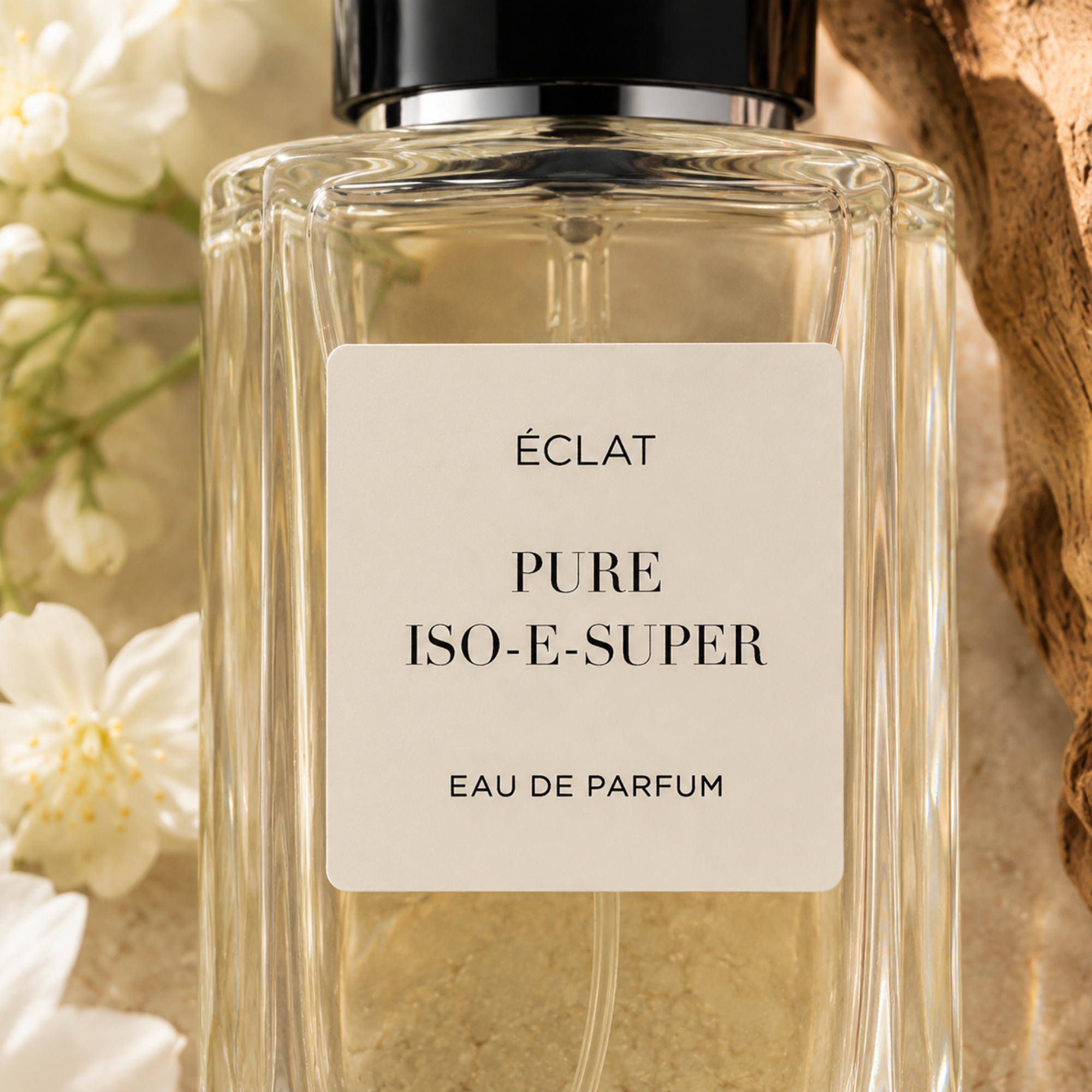 Un flacone in vetro sfaccettato di ÉCLAT Pure Iso-E-Super con tappo nero, circondato da fiori bianchi e legno strutturato.