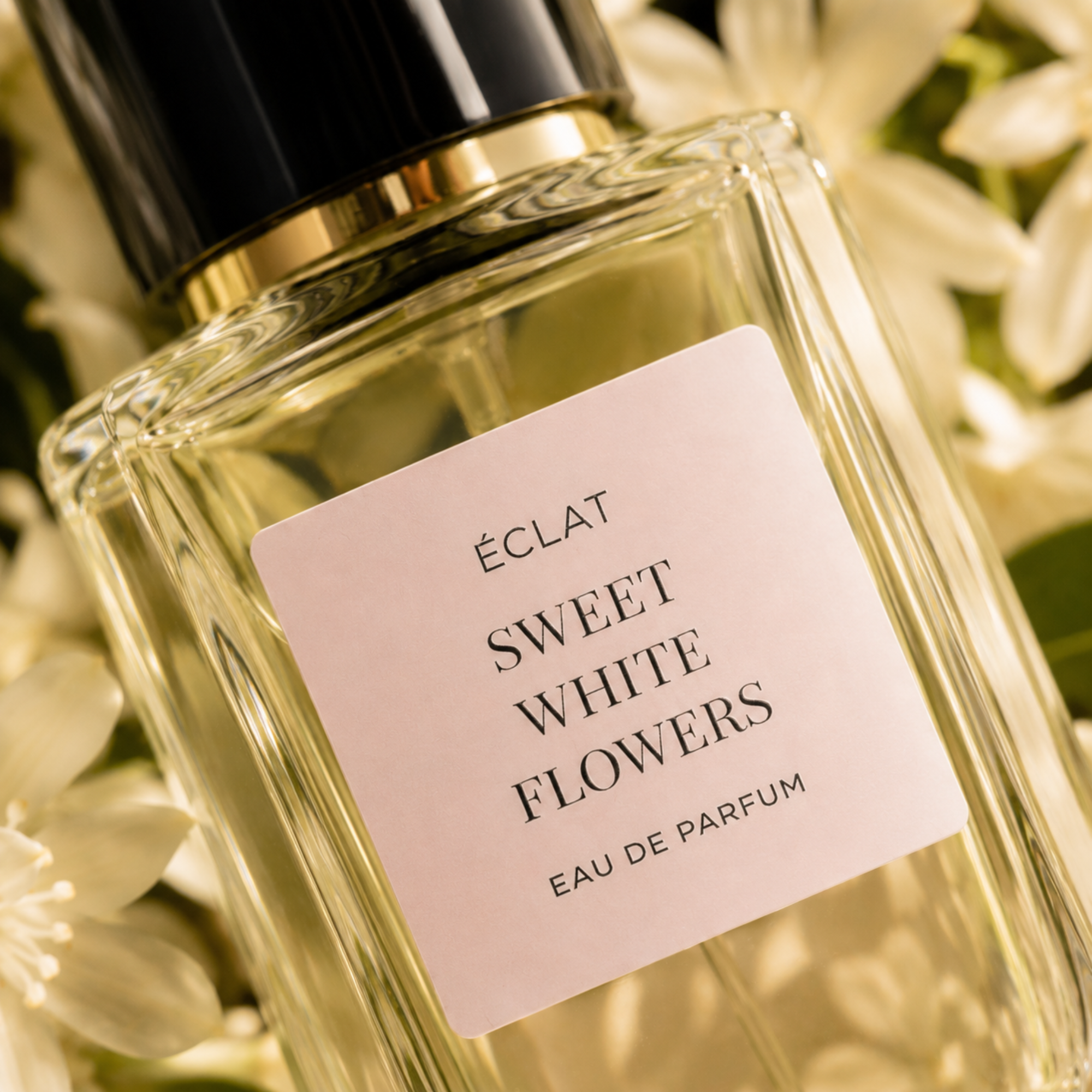 Un flacone in vetro sfaccettato di ÉCLAT Sweet White Flowers con etichetta rosa tenue e tappo nero e oro, circondato da fiori bianchi.