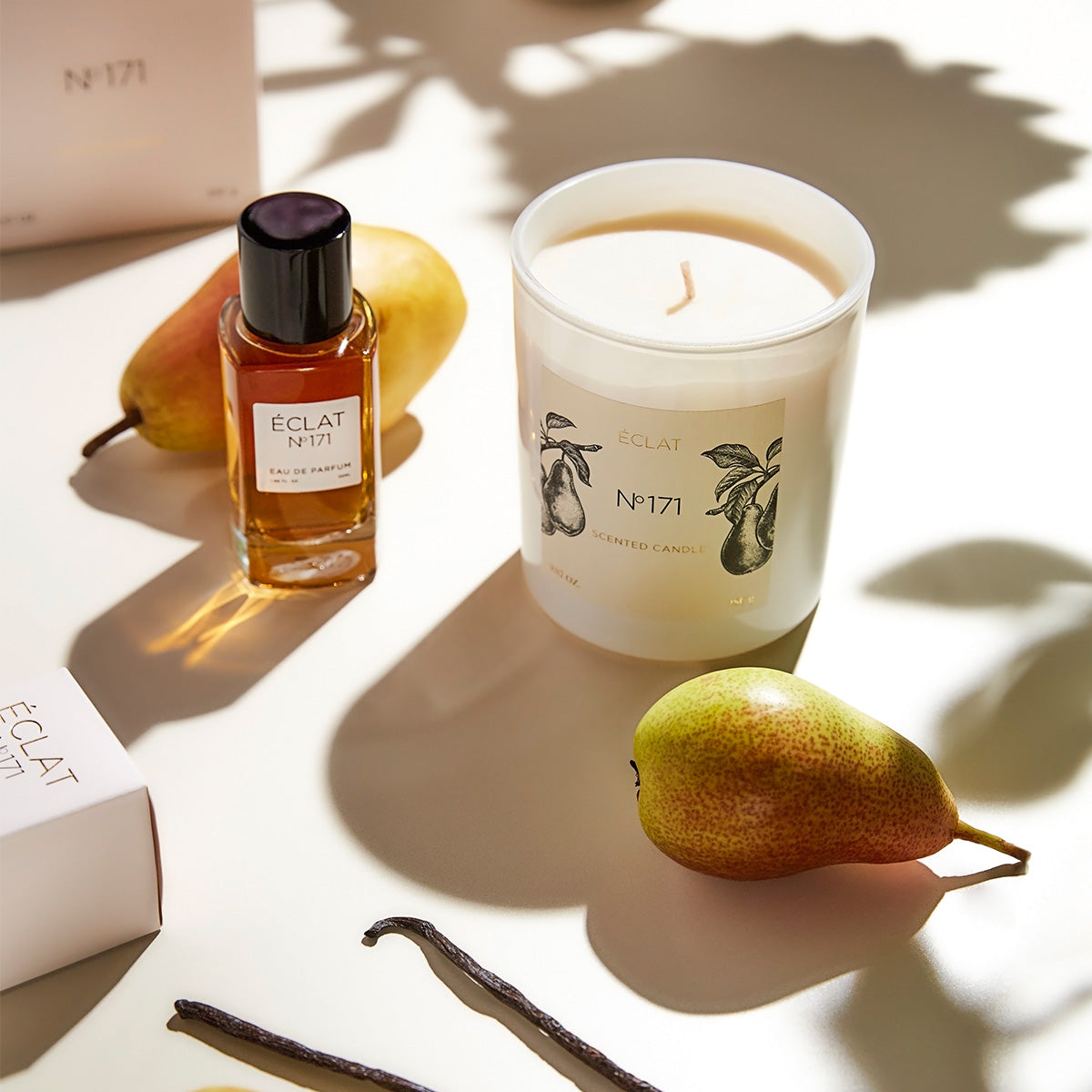 Flacone in vetro del profumo ÉCLAT Blackcurrant & Praline con tappo nero accanto a una elegante candela bianca e pera su sfondo chiaro.