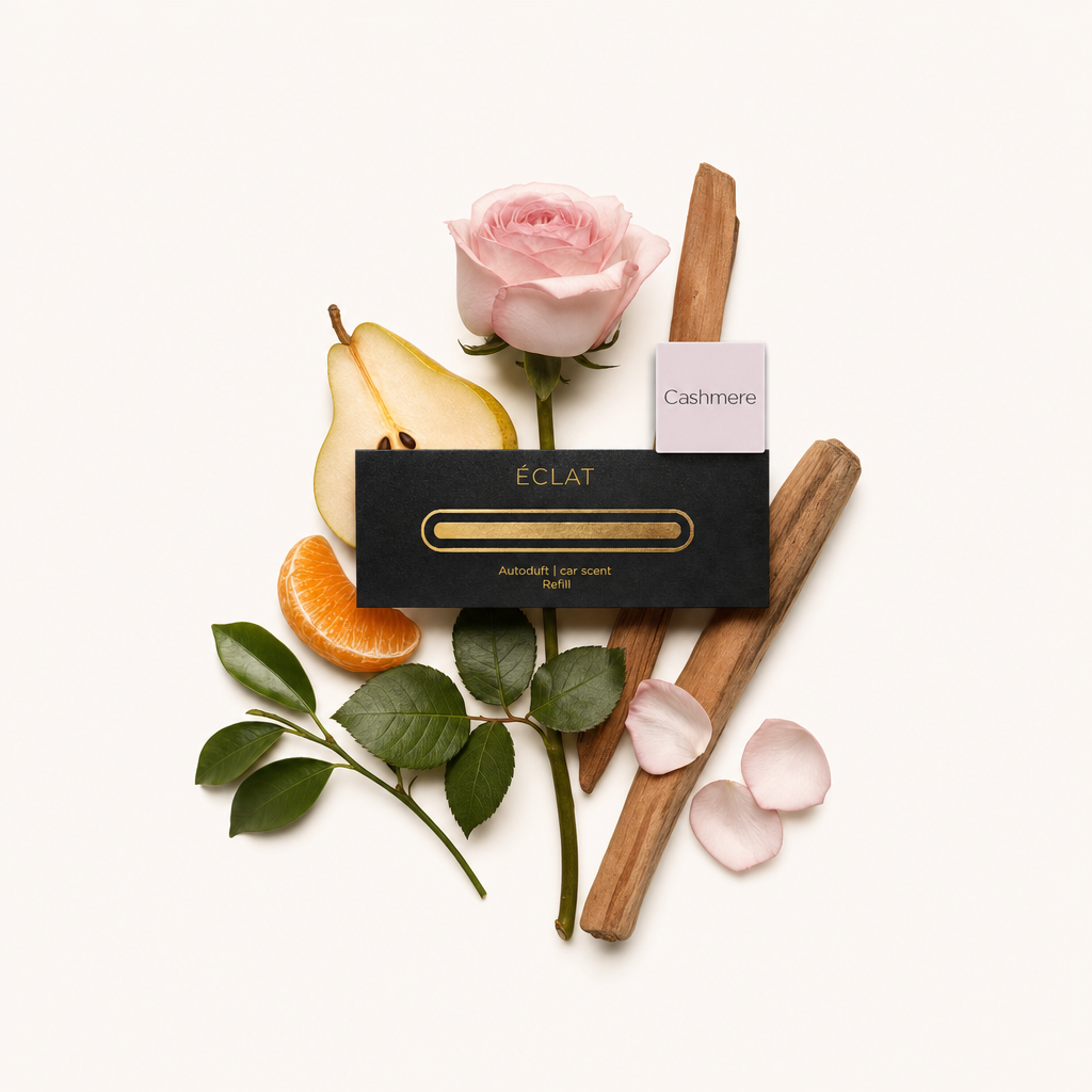 Un delicato arrangemento con una rosa rosa tenue, pera a fette, segmento di mandarino, due bastoncini di legno, foglie verdi e petali sparsi, accanto al tappo nero di ÉCLAT con etichetta 'Cashmere'.