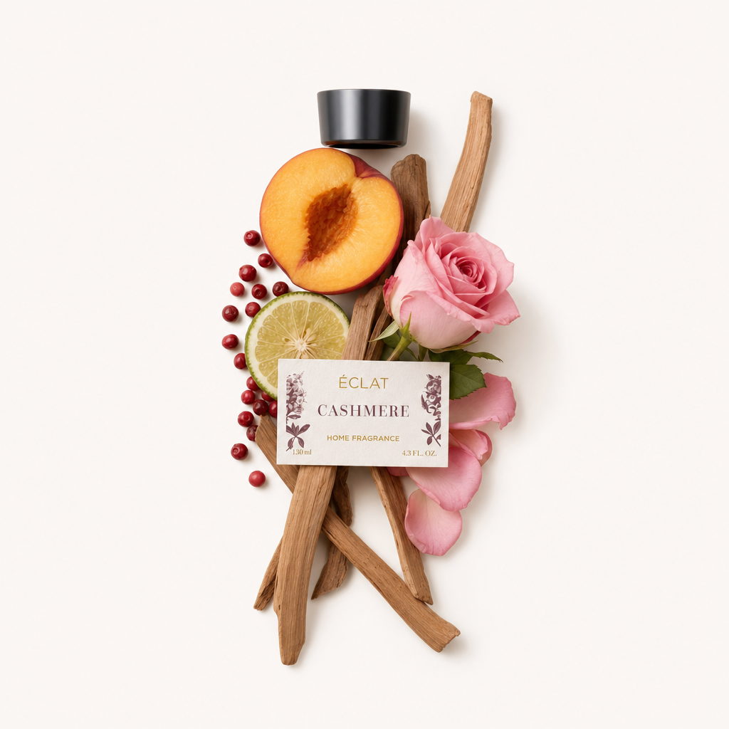Un arrangiamento di bastoncini di sandalo, una pesca dimezzata, una fetta di lime, rosa rosa e bacche rosse circonda un tappo nero etichettato ÉCLAT Cashmere Room Fragrance Sticks.