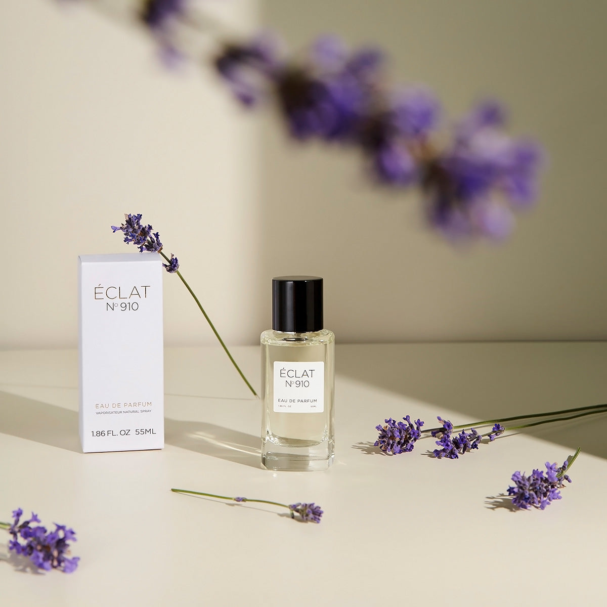 Profumo ÉCLAT Creamy White in un flacone di vetro trasparente con tappo nero, circondato da rametti di lavanda, accanto alla sua elegante scatola bianca.