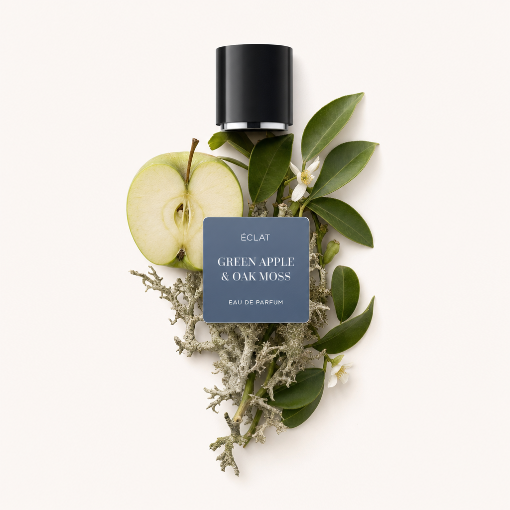 Una composizione di mezza mela verde, muschio di quercia e foglie verdi rigogliose si trova sotto un tappo nero con un'etichetta che presenta ÉCLAT Green Apple & Oak Moss.