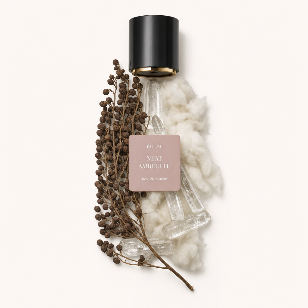 Un arrangiamento curato di semi marroni e morbido cotone bianco soffice accanto a strutture cristalline trasparenti. In alto si trova un tappo nero con etichetta beige che presenta ÉCLAT Neat Ambrette.