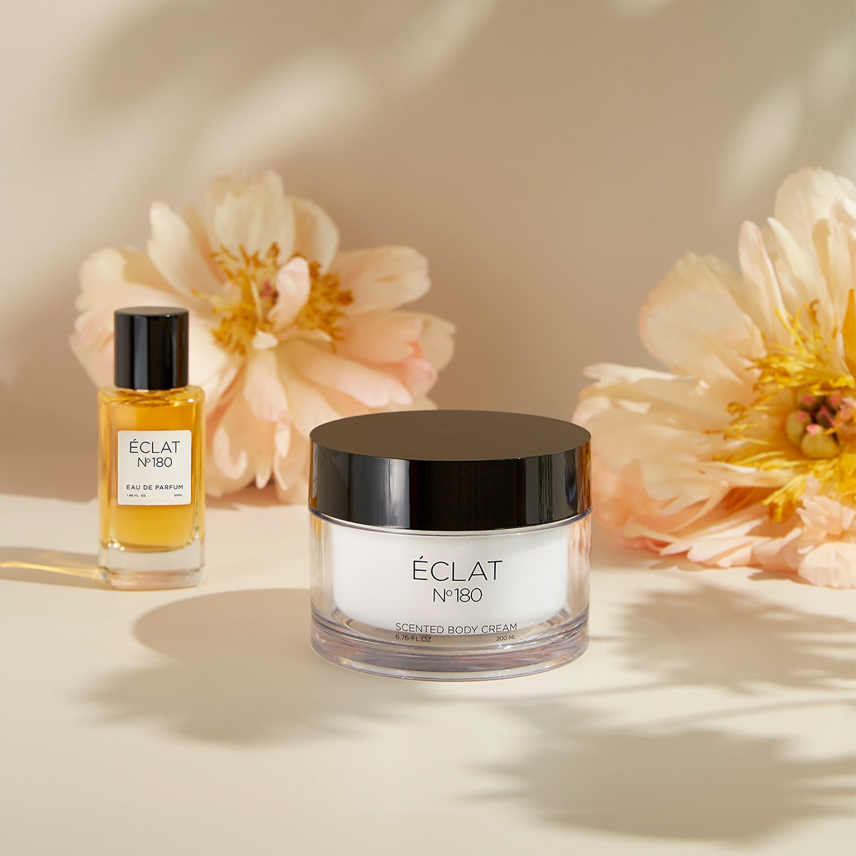 Set ÉCLAT Orchid & Patchouli con flacone di profumo in vetro trasparente e vasetto di crema con coperchio nero, circondato da fiori delicati.