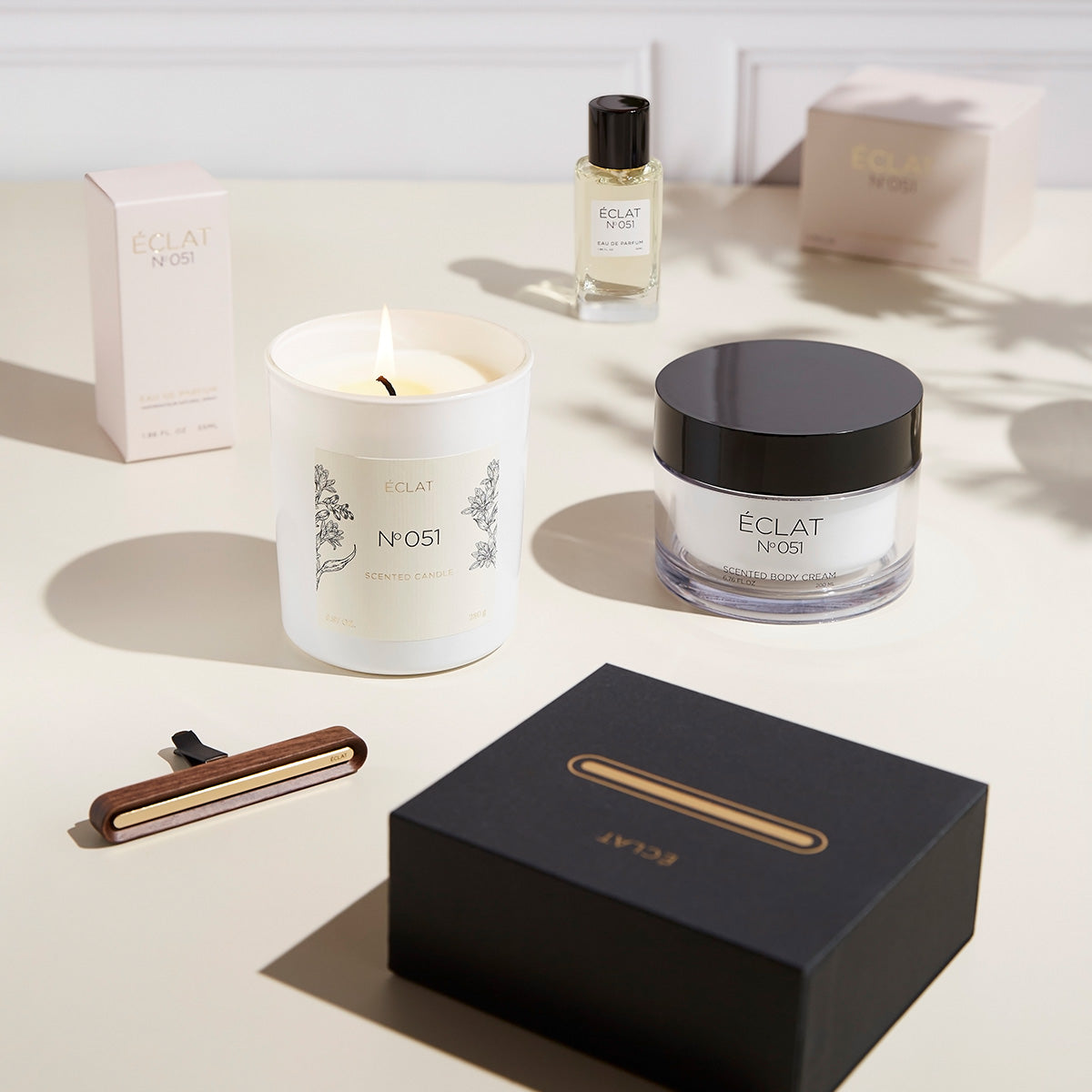 Una composizione elegante con una candela profumata accesa, un vasetto di vetro sfaccettato con coperchio scuro, una bottiglia di profumo e una scatola raffinata, che incarna ÉCLAT Oriental Almond.