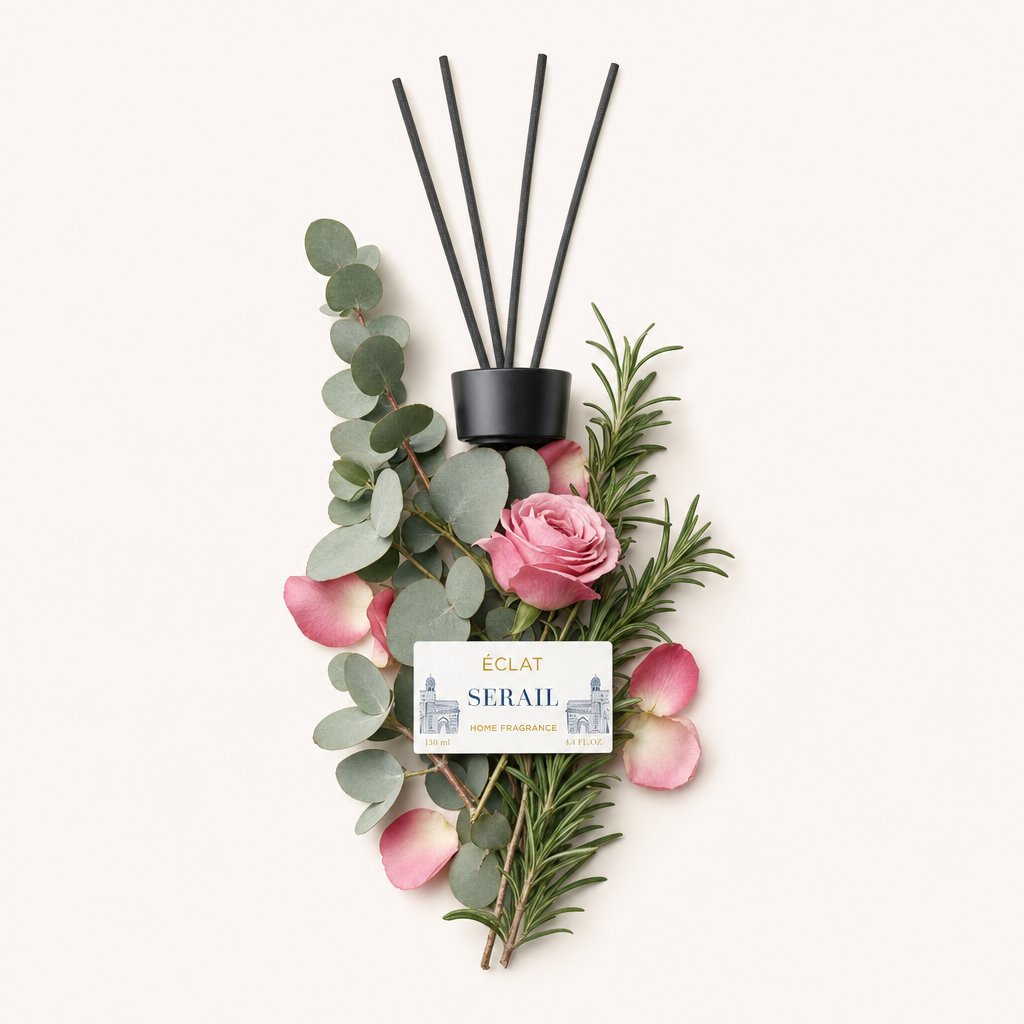 Un arrangiamento di foglie di eucalipto, rametti di rosmarino, petali di rosa rosa e una rosa fiorita circonda un tappo nero con etichetta per ÉCLAT Serail Room Fragrance Sticks.