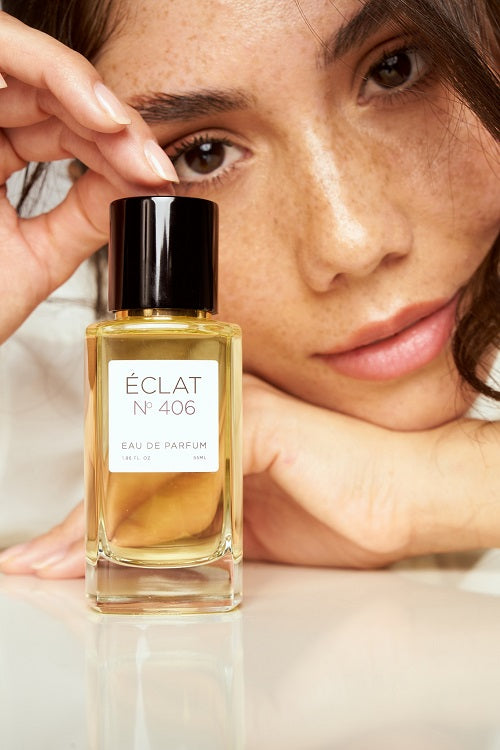 Un flacone di vetro sfaccettato di ÉCLAT N° 406 Eau de Parfum con tappo nero, davanti al volto di una persona.